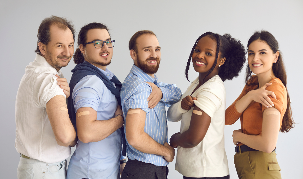 5 smiling people standing close together, showing off their bandaids from vaccination.