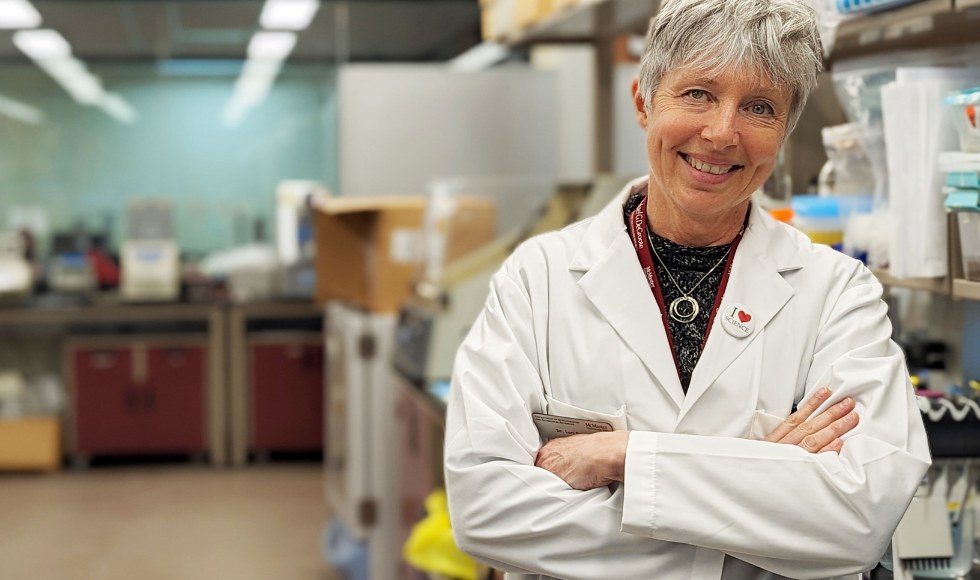 Lori Burrows standing in a laboratory, wearing a white lab coat and smiling at the camera