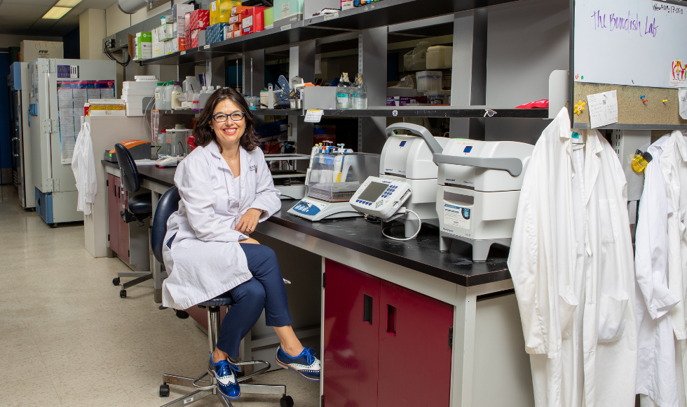 Smiling Dawn Bowdish wearing a lab coat and seated in an office chair at a desk in a lab full of equipment and lab coats.