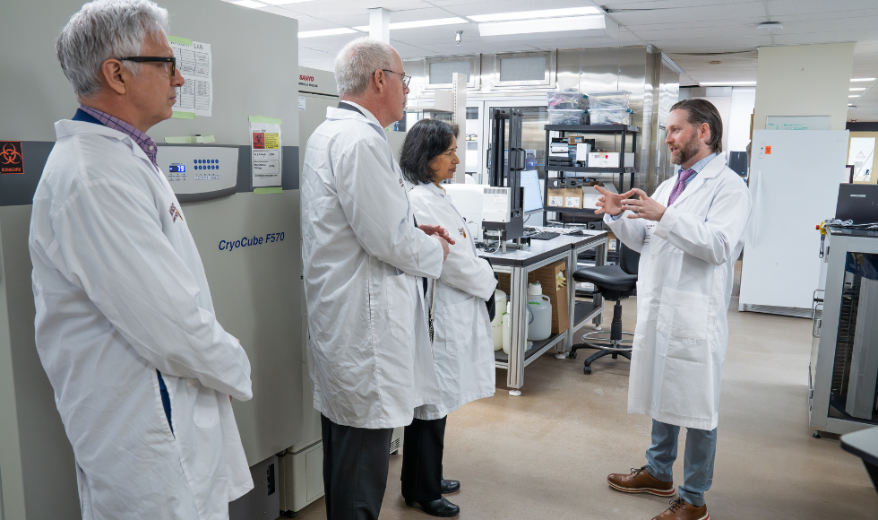 Professor Miller, in a lab coat, is gesturing at visitors, also in lab coats, inside a brightly lit lab.