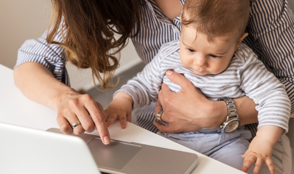 A baby in a woman's lap as they sit in front of a laptop computer. The adult's face is not in the frame.