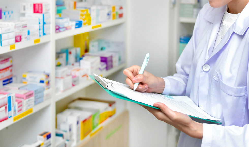 A pharmacist wearing a white lab coat writing on a pad of paper. There is a shelf of medications in the background.
