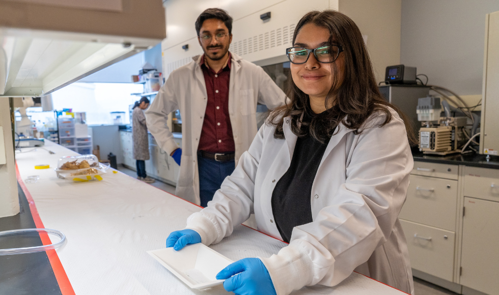 Two scientists in lab coats smiling at the camera in a lab. There is a tray with food in it on the counter.