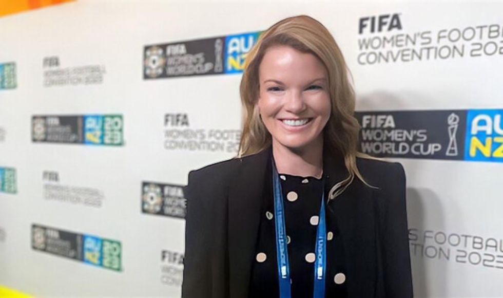 Sinead Dufour smiling at the camera. Behind her, there is a backdrop branded with the FIFA Women’s World Cup logo.