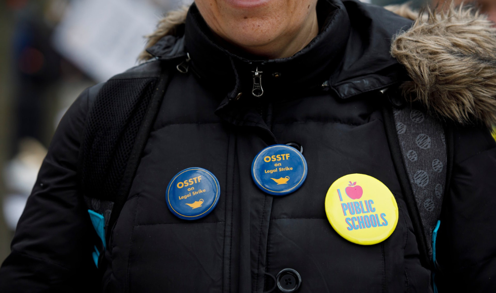 A person wearing a black winter coat with buttons pinned on it that read, ‘OSSTF on Legal Strike’ and ‘I [image of an apple] public schools.’