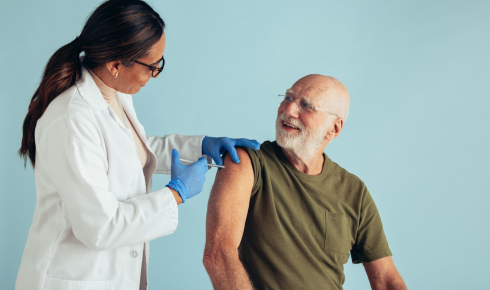 A smiling person in a lab coat is giving a smiling older gentleman an injection.