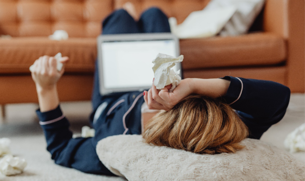 Seen from behind, person in PJs lying on the floor with a screen propped up on their belly, clutching a tissue and surrounded by used tissues.