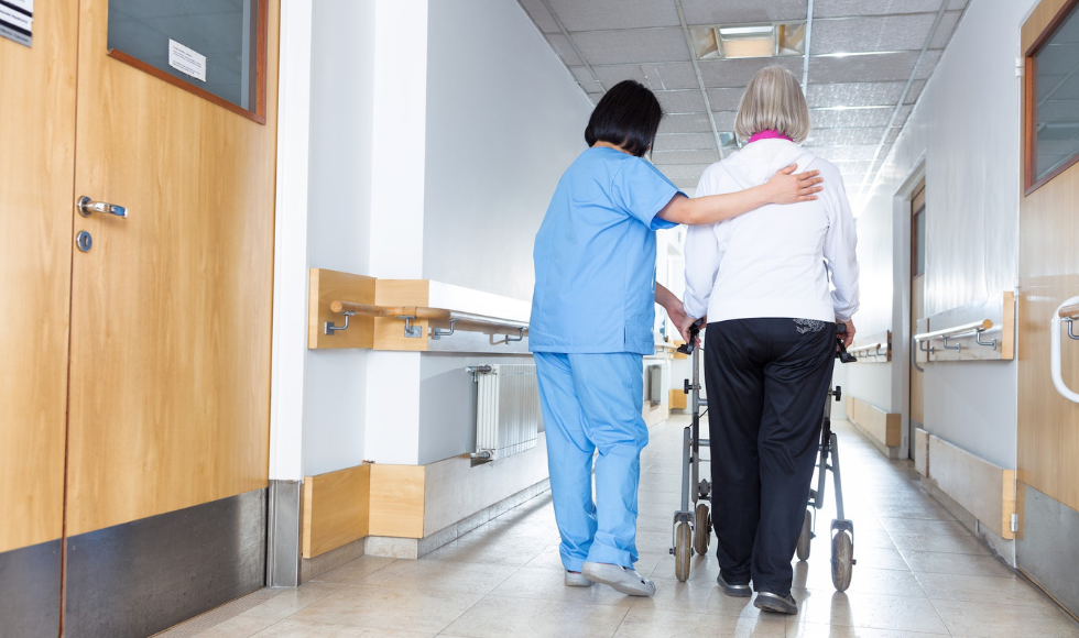 Seen from behind: An older adult using a walker alongside a care worker in scrubs in the hallway of a building