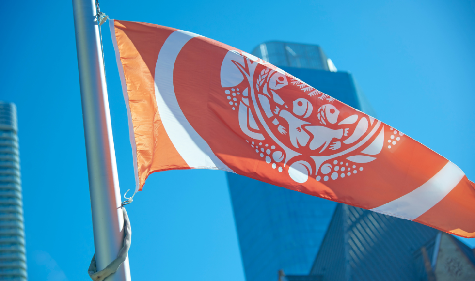An orange Survivor's Flag in the foreground with skyscrapers against a blue sky behind it.