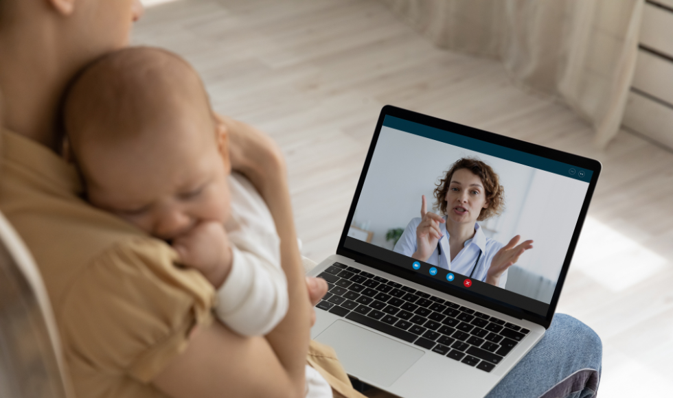 A doctor is on a computer screen balanced on the lap of an adult holding a napping baby.