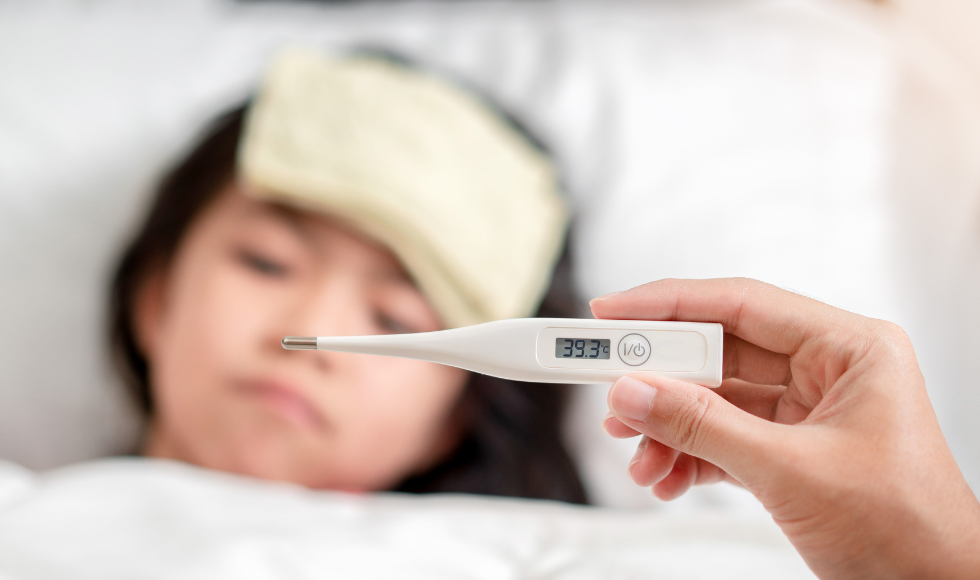 hand holding a digital thermometer showing a fever in the foreground. In the background is a sleeping child with a compress on their forehead.