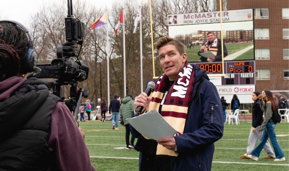 Image of Robert Cockcroft on the field doing a live report, with a microphone in his hand and a large video camera pointed at him