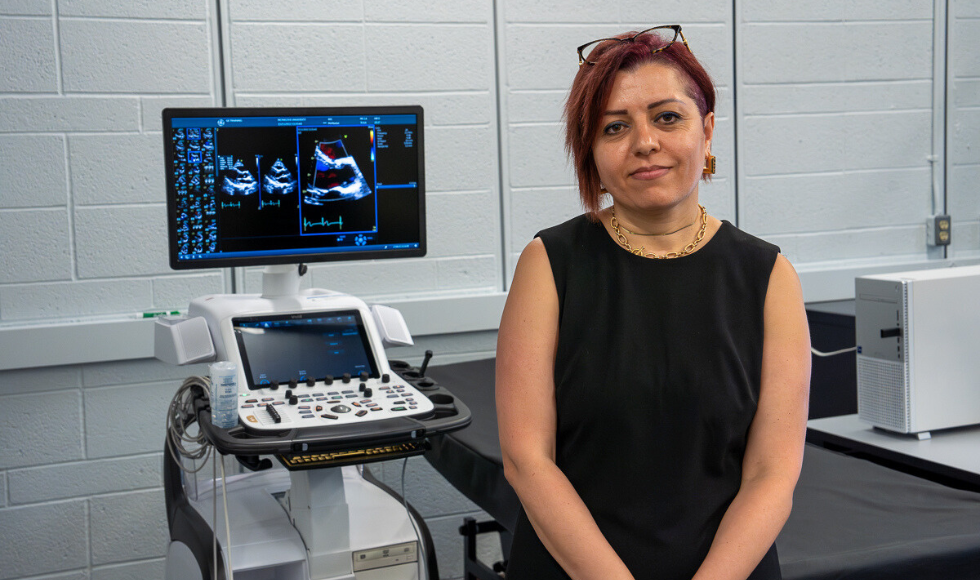 Waist-up shot of Zahra Motamed wearing a black sleeveless top with glasses on top of her head, in a lab, with a computer showing diagnostic imaging in the background.