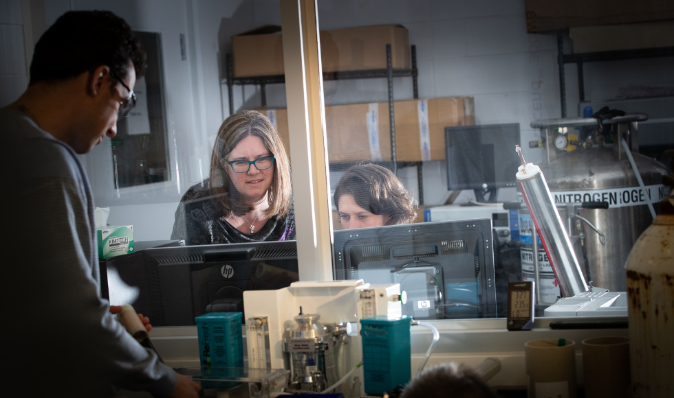 Gillian Goward and a colleague are seen through a glass pane in an industrial lab setting, looking at a computer while surrounded by equipment. Another colleage is off to the left in the foreground.