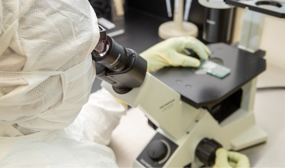 Over the shoulder look at a person in full head-to-toe protective lab gear looking into a microscope