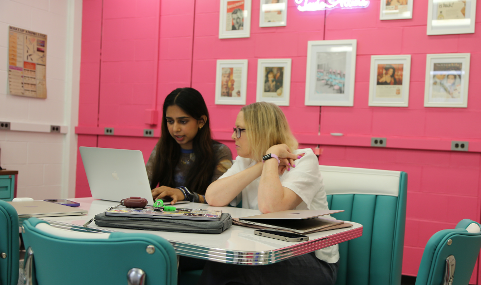 Siya Sood and Fiona McNeill work at a laptop on a Tiffany blue diner table with matching chairs, against the backdrop of a hot pink wall. It looks like they're in a diner.