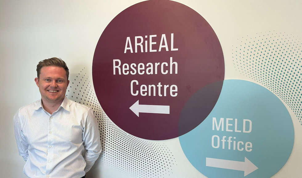 Daniel Schmidtke smiling at the camera while standing beside two signs that read, 'ARiEAL Research Centre' and 'MELD Office.'