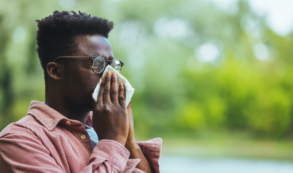 A man blowing his nose. There is greenery out of focus behind him.