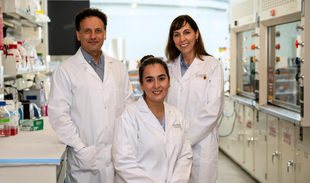 Two researchers stand on either side of a third seated researcher, all smiling and wearing lab coats, inside a lab.