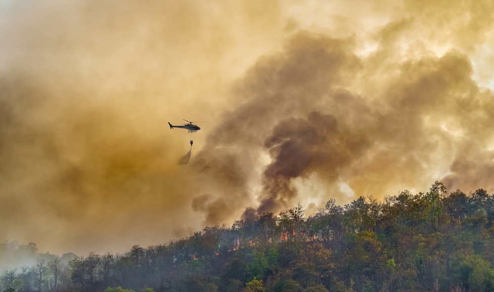 A helicopter flies over a wildfire. The sky is filled with smoke and a yellow haze.