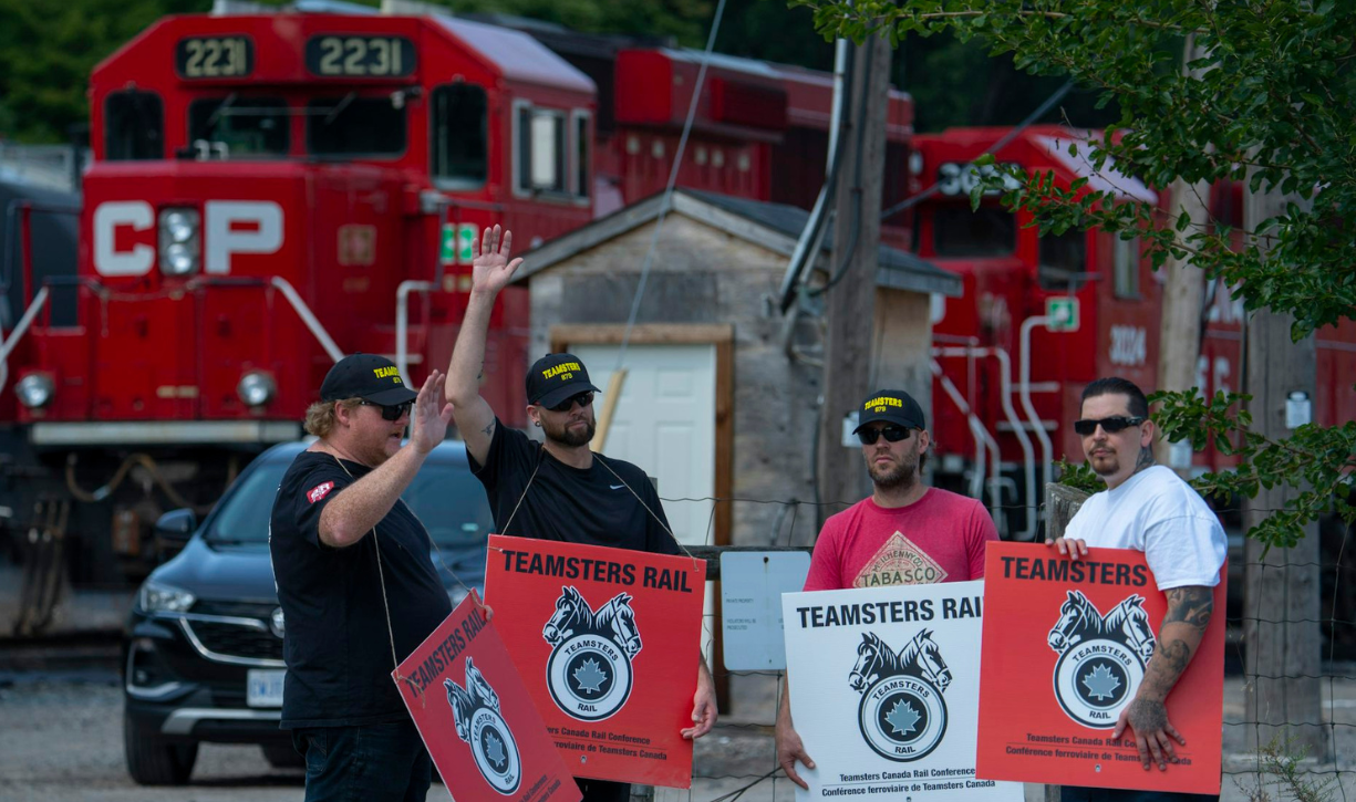 A group of men in t-shirts and baseball caps holding signs that say 'Teamsters Rail' stand in front of a stationary freight train