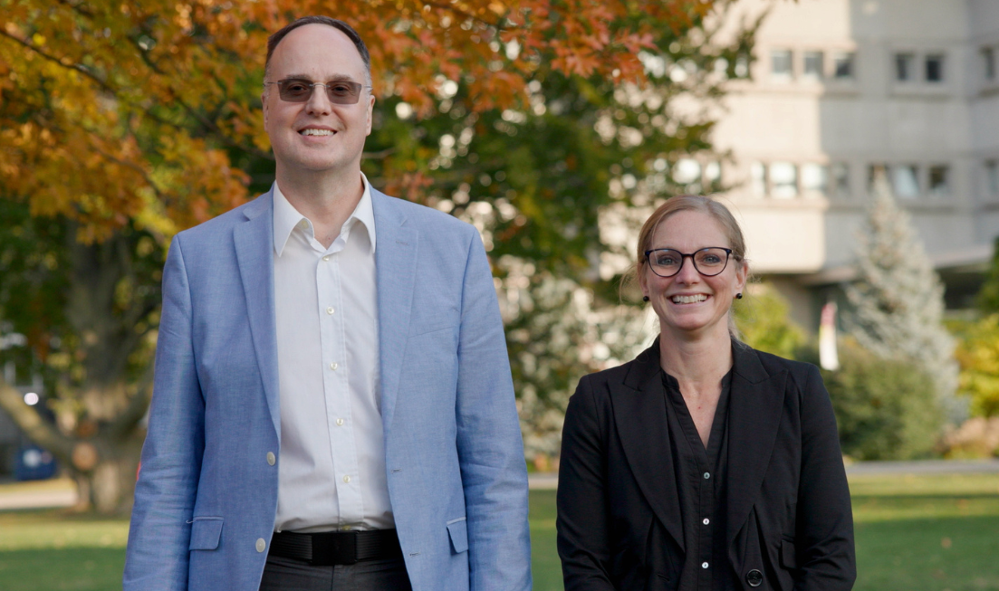 Two smiling people stand side by side outdoors in the fall, with trees in the background.