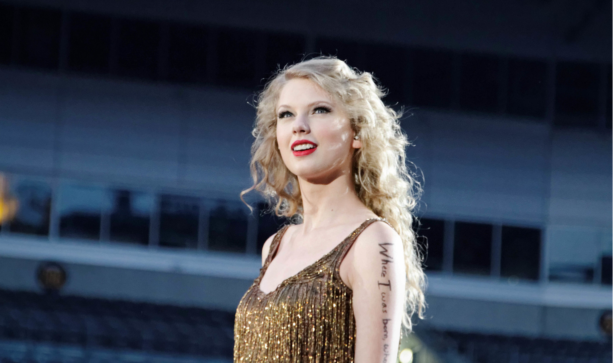 Taylor swift in concert wearing a glitzy metallic tank dress with writing on her arm.