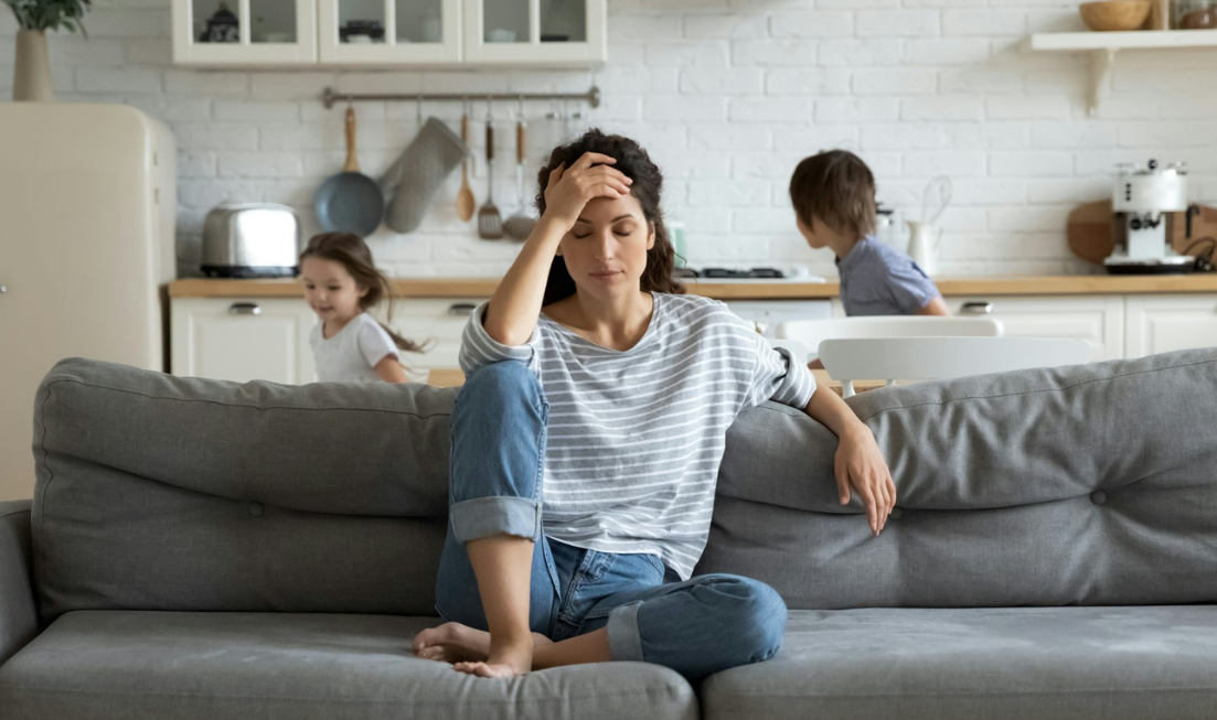 A woman sits on a couch in a tired/exasperated posture while kids run around in a blur of motion behind her.