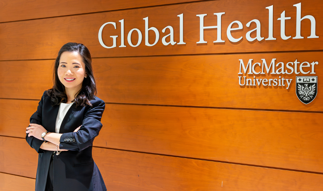 A smiling person in a suit standing with their arms crossed in front of the McMaster Global Health sign in the Faculty of Health Sciences.