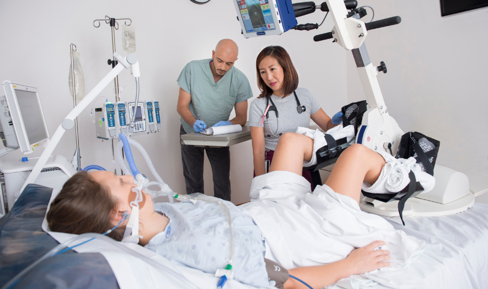 A person lying in a hospital bed with their feet in a set of bicycle pedals that are attached to a device that has a monitoring screen attached to it. There is a woman with a stethoscope hanging over her shoulders at the side of the bed and another person in the background taking notes.