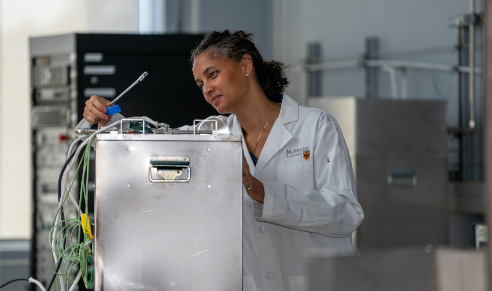 Keena Trowell wearing a white lab coat that has the McMaster University logo on it while working over a piece of lab equipment.