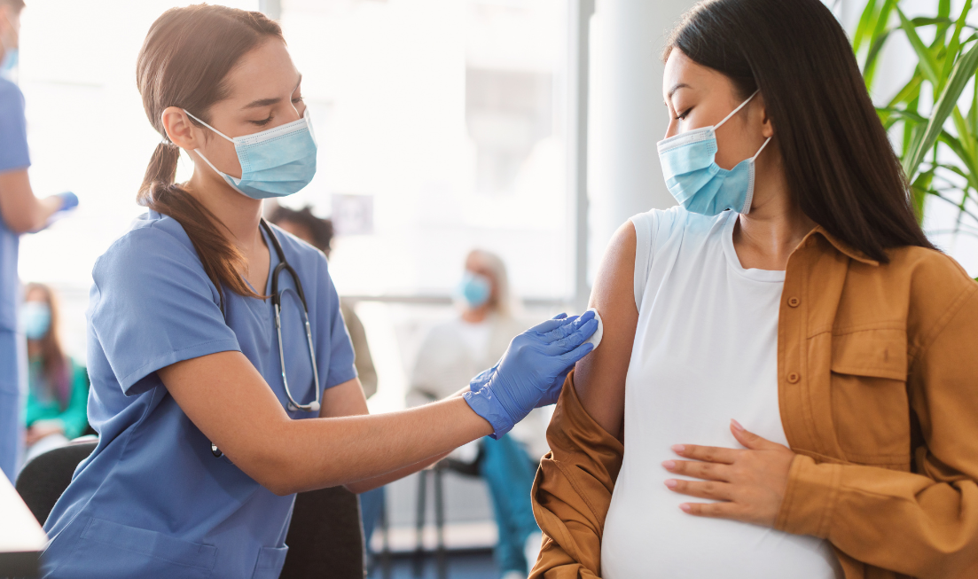 A person in a mask and scrubs swabs the upper arm of a masked pregnant person who is getting vaccinated.