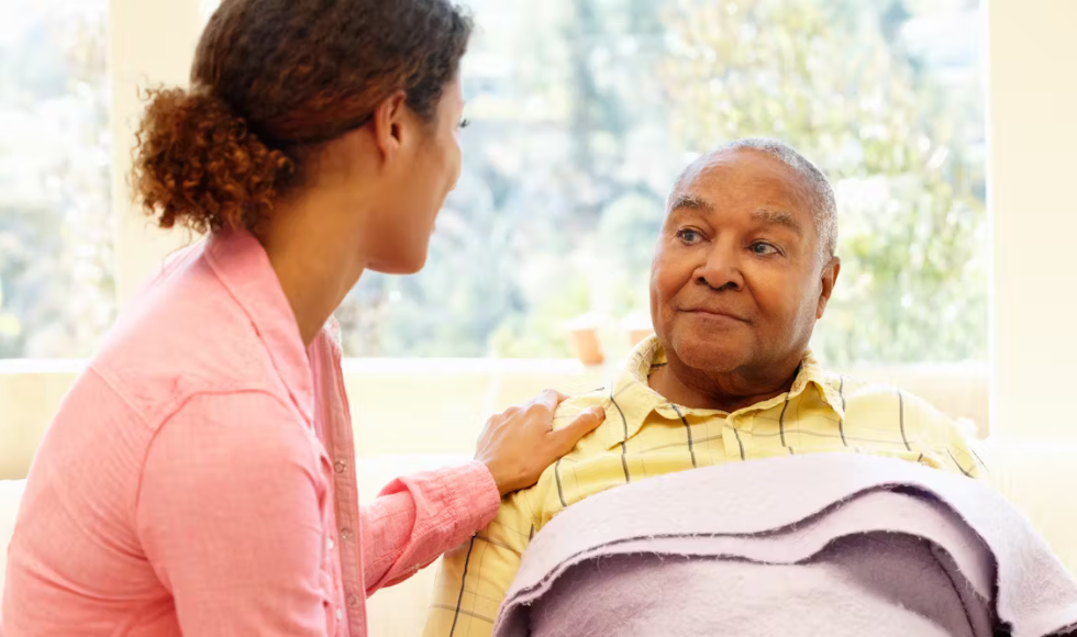 A person putting their hand on the shoulder of a person who is seated with a blanket on them. The two people are looking directly at each other.