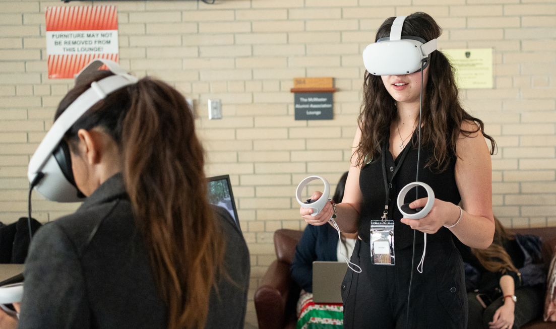 Two people with long dark hair wearing virtual reality headsets and holding controllers in each hand. One person seen from behind, one is facing us.