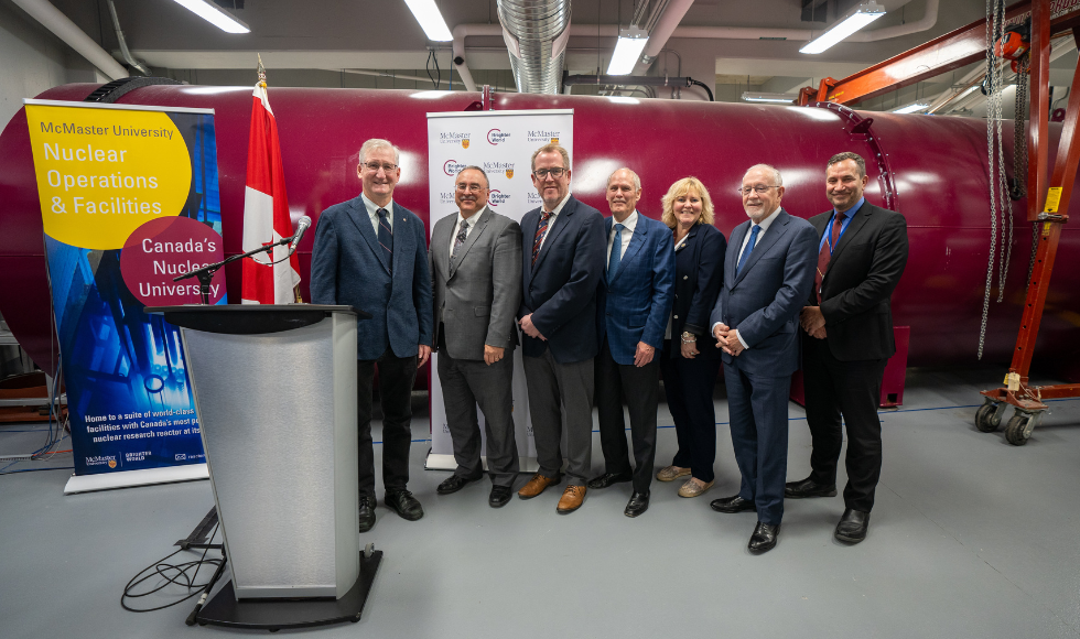 Seven people lined up in front of a maroon neutron beam smiling at the camera. There is a podium with a microphone and two banners with McMaster University branding on them.