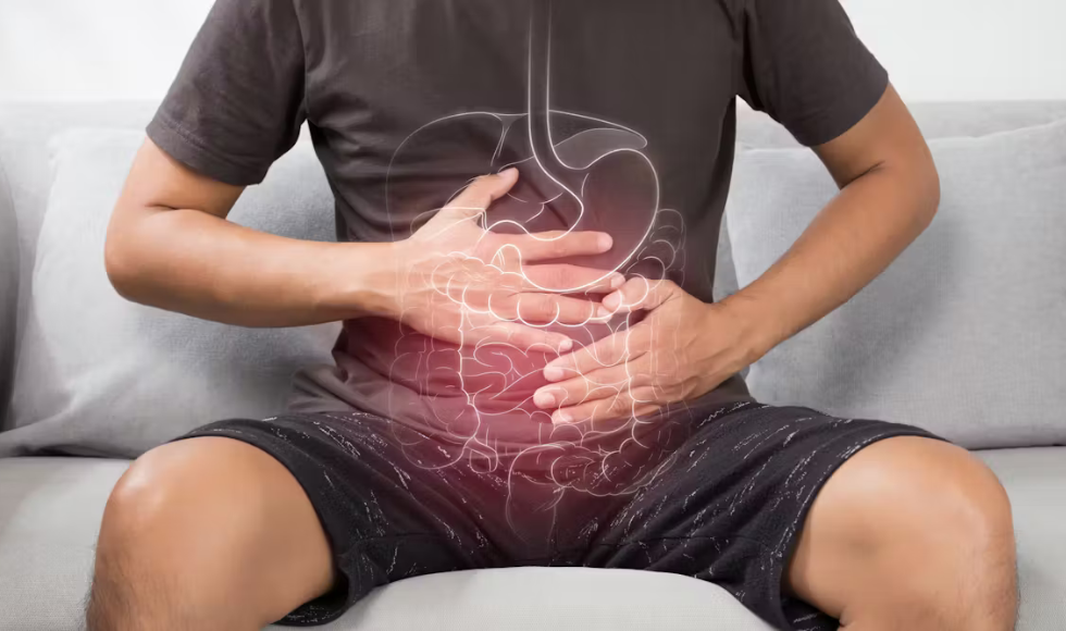 A photo showing the midsection of person sitting on a grey couch in a black t-shirt and shorts. Their hands are resting on their abdomen and there is a graphic design of internal organs overlaid over the person’s stomach area.