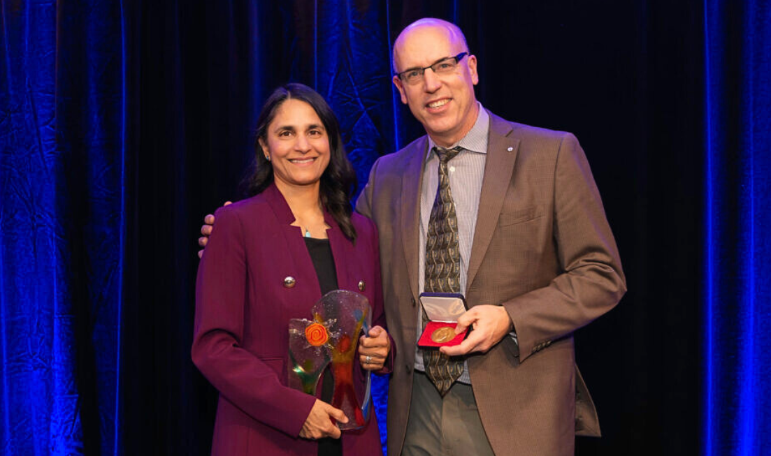 Sonia Anand and a man in a brown suit stand together, smiling and holding an award and a medal.