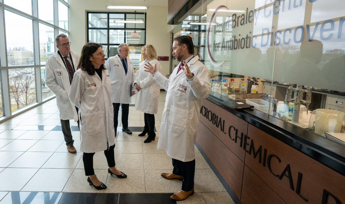 Five people in lab coats standing and talking in a wide hallway between a wall of windows and office space labelled The David Braley Centre for Antibiotic Discovery.