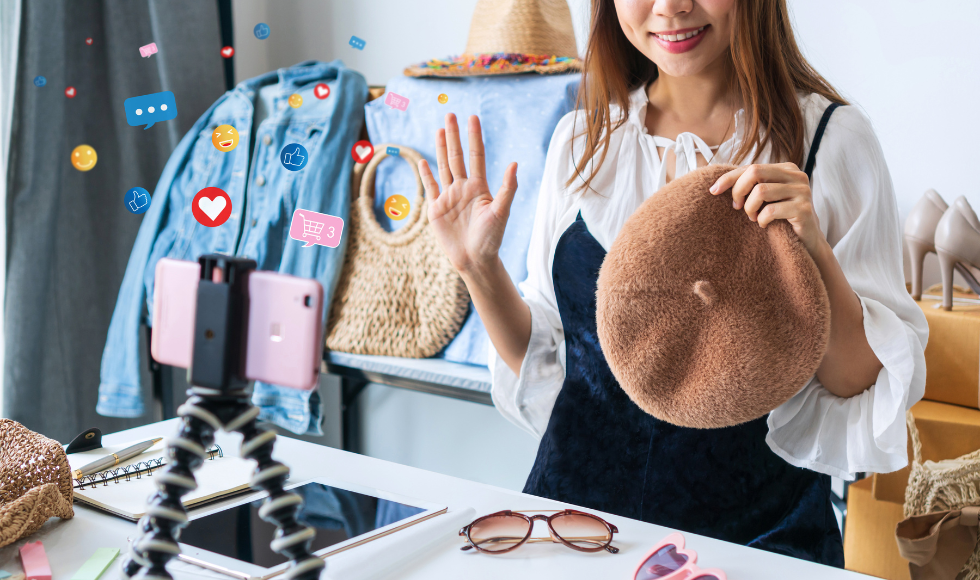 A woman smiling and holding up a hat while standing in front of a cellphone that is attached to a stand that is sitting on top of a table. There are graphic overlays of emojis around the cellphone.