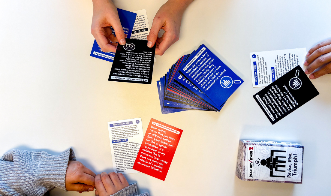 Seen from directly above, three sets of hands holding or displaying red, blue, white and black cards from the game Dear Reviewer 2.