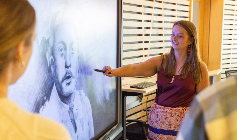 Tekenikhon Doreen, seen over the shoulders of two people, points to a portrait of Deskaheh Levi General on a big screen.