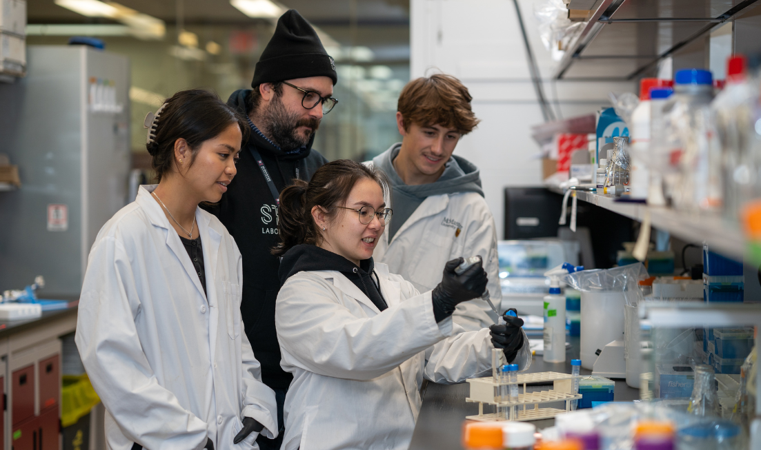 Watched by a professor in a toque and hoodie and two grad students in lab coats, a grad student works with a pipette and a beaker in a lab at McMaster.