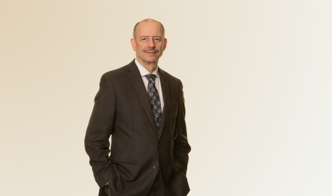 A man in a dark brown suit, light-coloured shirt and dark tie standing against a light background with his hands in his pocket