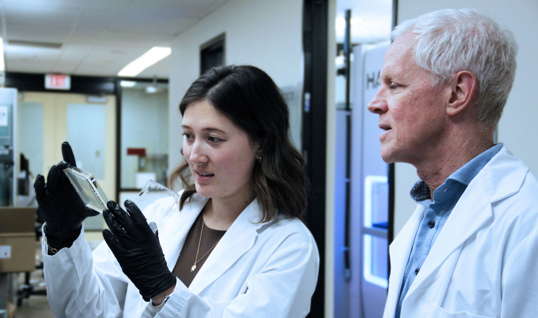 Researchers Megan Tu and Eric Brown, both wearing lab coats, are in a lab, looking at a sample that Tu is holding in her gloved hands.