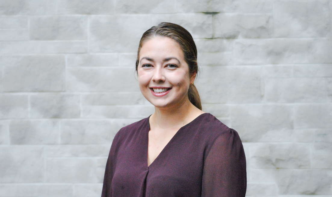 Head and shoulders of Rebecca Correia, smiling and wearing a dark blouse against the backdrop of a grey stone wall.