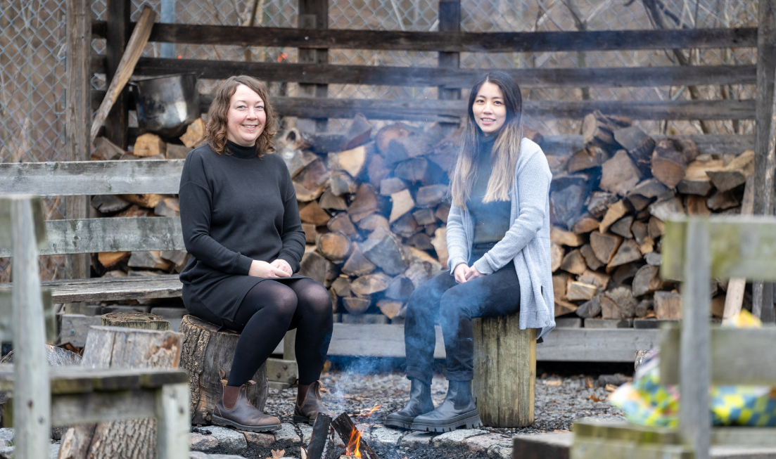 Researchers Sarah Styler, and Iris Chan sitting on wooden benches at a small bonfire, in front of a few cords of stacked firewood.