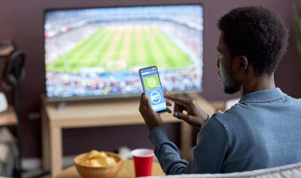 A person looks at a cellphone. In the background there is a TV displaying a sporting event.