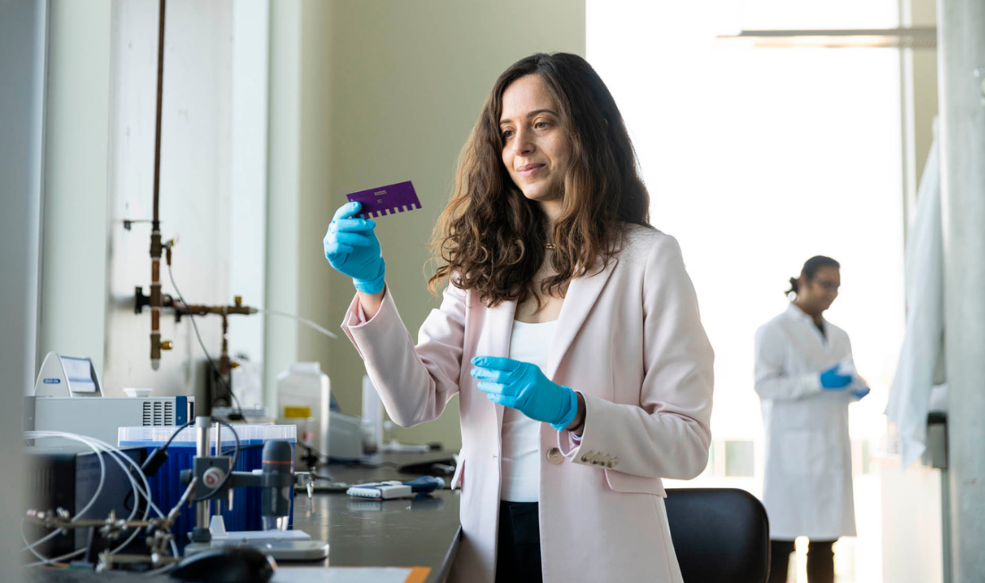 Prof Leyla Soleymani in a lab, looking at a rectangular device she's holidng in her gloved hand. In the background (soft focus) a graduate student in a lab coat.