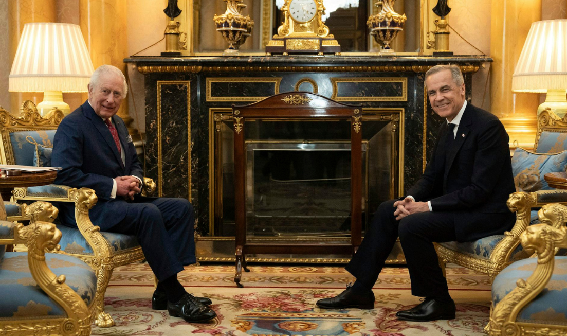 An older man with grey hair sits in an ornate room across from another man with greying hair. Both smile at the camera.