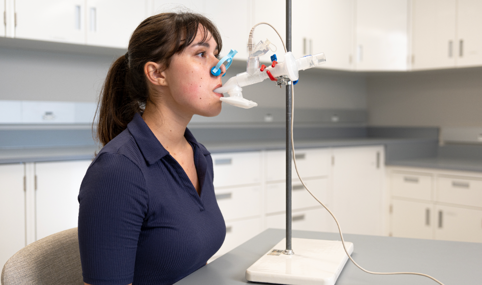 A woman sits at a table in a clinical room. On the table is a medical apparatus on a stand, with a tube extended from it that she is holding in her mouth. She has a light blue clamp over her nose.